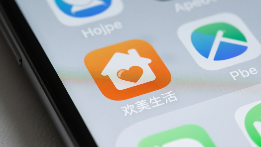 一张欢美生活APP在手机桌面的图标特写,图标以橙色为底,中间有白色房子和心形图案,手机屏幕背景为简约的浅灰色壁纸,周围有其他应用图标虚化,突出欢美生活APP图标,整体画质清晰,干净无多余元素。