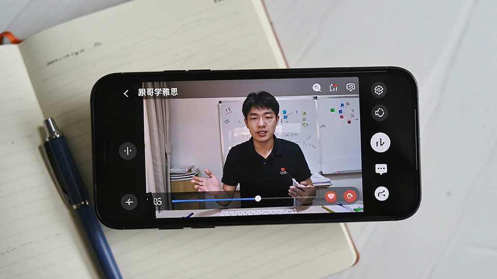 手机横屏使用“跟哥学雅思”观看直播课的实拍图,主体为手机屏幕显示外教授课画面,旁边有笔记本和笔,布局为俯拍视角,风格自然生活化,细节包括屏幕上的进度条和互动按钮,画质清晰干净