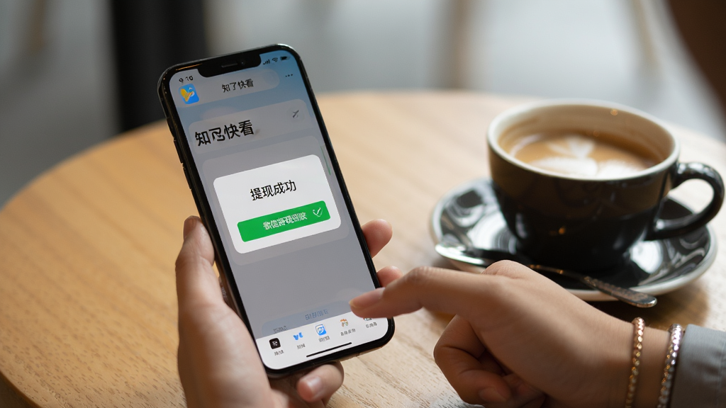 一张用户使用手机操作“知了快看”APP的实拍图,手机屏幕显示“提现成功”的弹窗,弹窗上写着“微信提现到账”,手机放在木质桌面上,旁边有一杯咖啡,背景虚化,整体氛围轻松,画质干净,突出实用感。