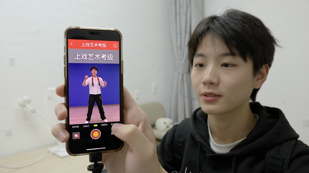 考生使用手机进行上戏艺术考级考试的场景预览图,画面中一位考生面对手机摄像头进行才艺展示,背景为简洁的室内环境,手机屏幕显示上戏艺术考级APP的录像界面,整体画面明亮清晰,采用纪实摄影风格,突出真实感与实用性