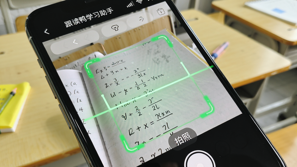 手机屏幕特写显示“跟读鸭学习助手”的拍照批改页面,摄像头对准写满数学题的作业本,屏幕上有绿色的识别框和“拍照”按钮,周围有动态的扫描线,背景是简洁的课桌,整体色调明亮,构图清晰,画质干净,无多余装饰。