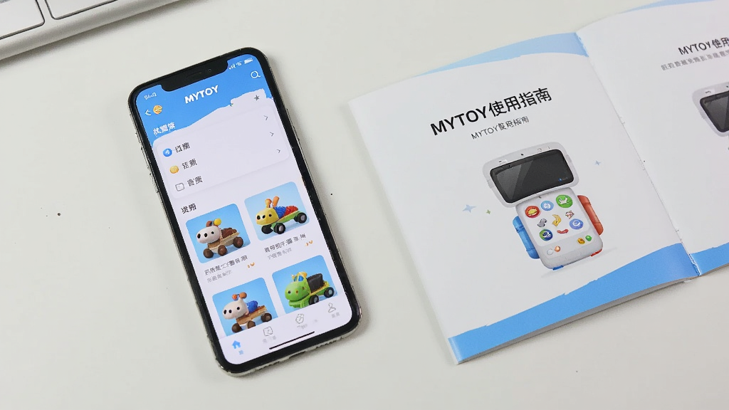 主体为一部手机平放在桌面上,屏幕显示MYTOY应用的主界面,界面中有分类列表和玩具卡片,手机旁边有一本打开的操作手册,手册上印有“MYTOY使用指南”字样,背景为简洁的办公桌,布局为俯视角度,排版清晰,风格为实拍风格,光线明亮,细节丰富,画质干净。