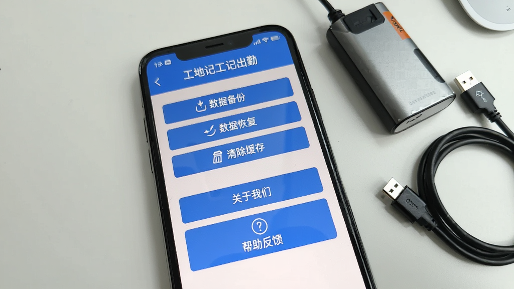 一张手机截图,展示“工地记工记出勤”的设置页面,页面顶部有“数据备份”、“数据恢复”、“清除缓存”等选项,下方有“关于我们”和“帮助反馈”按钮。背景为简洁的桌面,手机旁边放有移动电源和数据线,画面清晰,干净无多余元素。