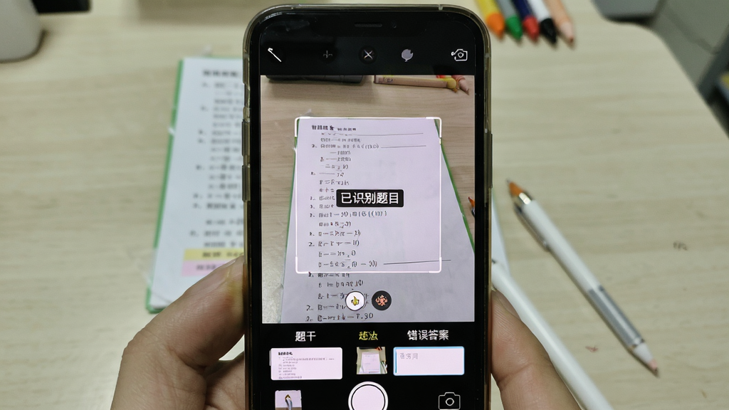 展示“错题本”软件拍照录入功能的预览图,主体为手机相机取景界面,对准桌面上的数学试卷,屏幕上有矩形取景框和“已识别题目”的提示标签。布局为第一视角手持手机拍摄,风格写实,细节包括界面下方的“题干”、“错误答案”输入栏缩略图,画质清晰,画面干净,突出功能便捷性。