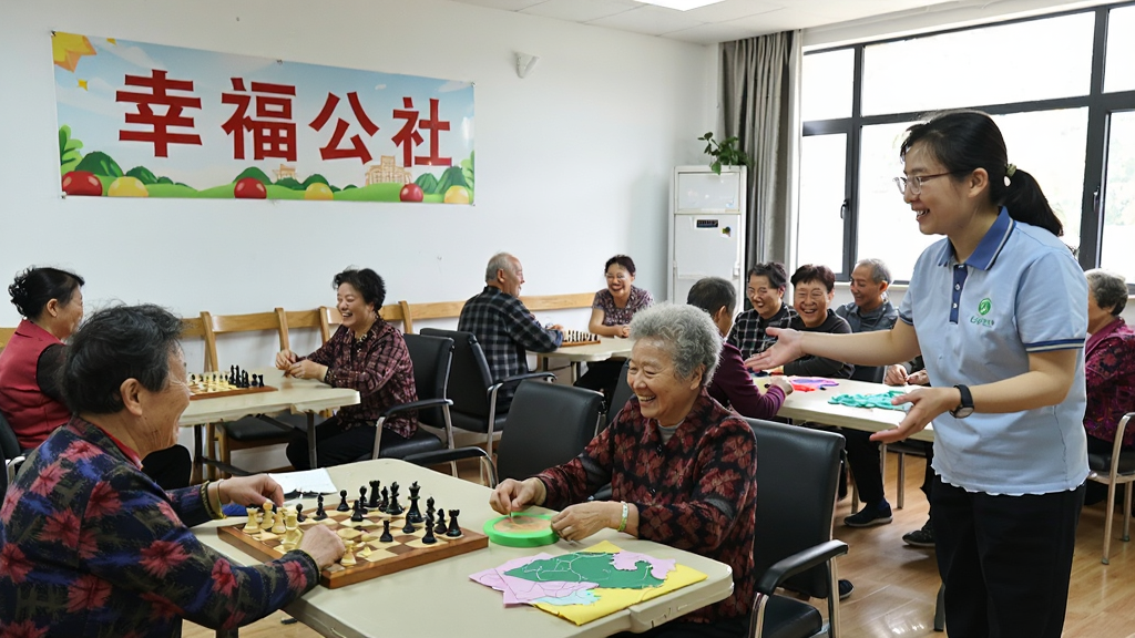 心智青幸福公社线下活动场景,一群中老年人在社区活动中心内下棋、聊天、做手工,墙上挂着“幸福公社”标识,氛围欢乐融洽,一位社长模样的工作人员正在热情引导,整体风格写实自然,光线明亮,画质清晰,无多余元素