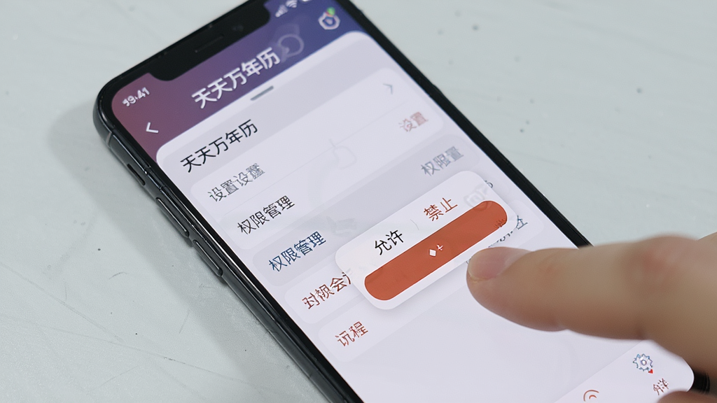 主体:一部手机正在运行天天万年历,屏幕上显示设置菜单和权限管理选项。布局:手机斜放,背景为浅灰色桌面。排版:界面清晰,菜单项突出。风格:实用教程风格。细节:手指点击“权限管理”选项,屏幕上弹出允许/禁止对话框。画质:干净,无多余元素。